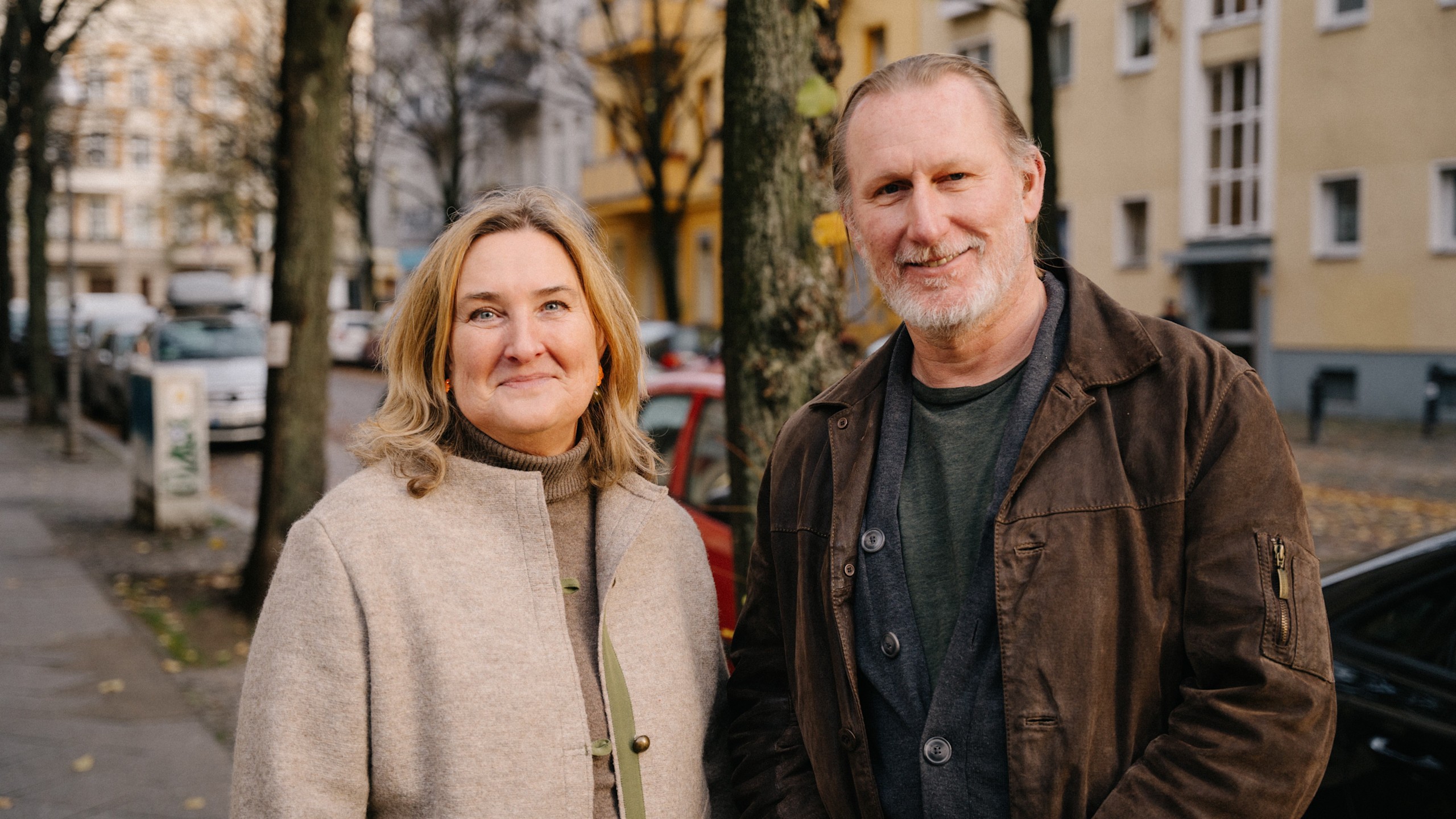 Schwerhörigkeit Portrait Paar 16:9 Porträt einer blonden Frau im beigen Mantel und eines Mannes mit grauem Bart und brauner Jacke. Beide lächeln freundlich vor einer unscharfen urbanen Kulisse mit Bäumen und Häusern.