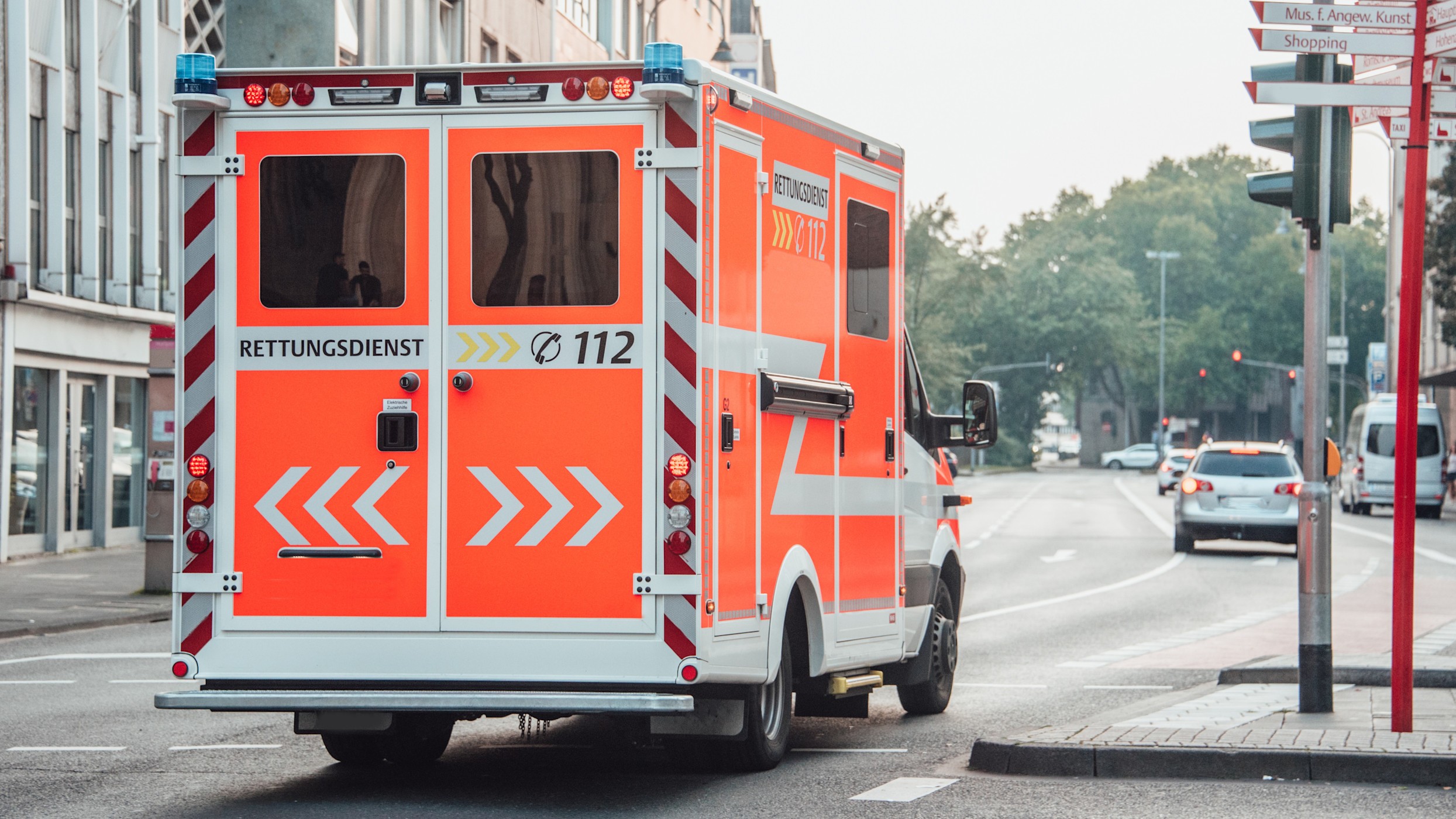 16:9 Header Einsatzkröfte Das Bild zeigt einen orangefarbenen Rettungswagen, der durch eine Stadt fährt. Es ist Tag, und andere Autos sind auf der Straße unterwegs.