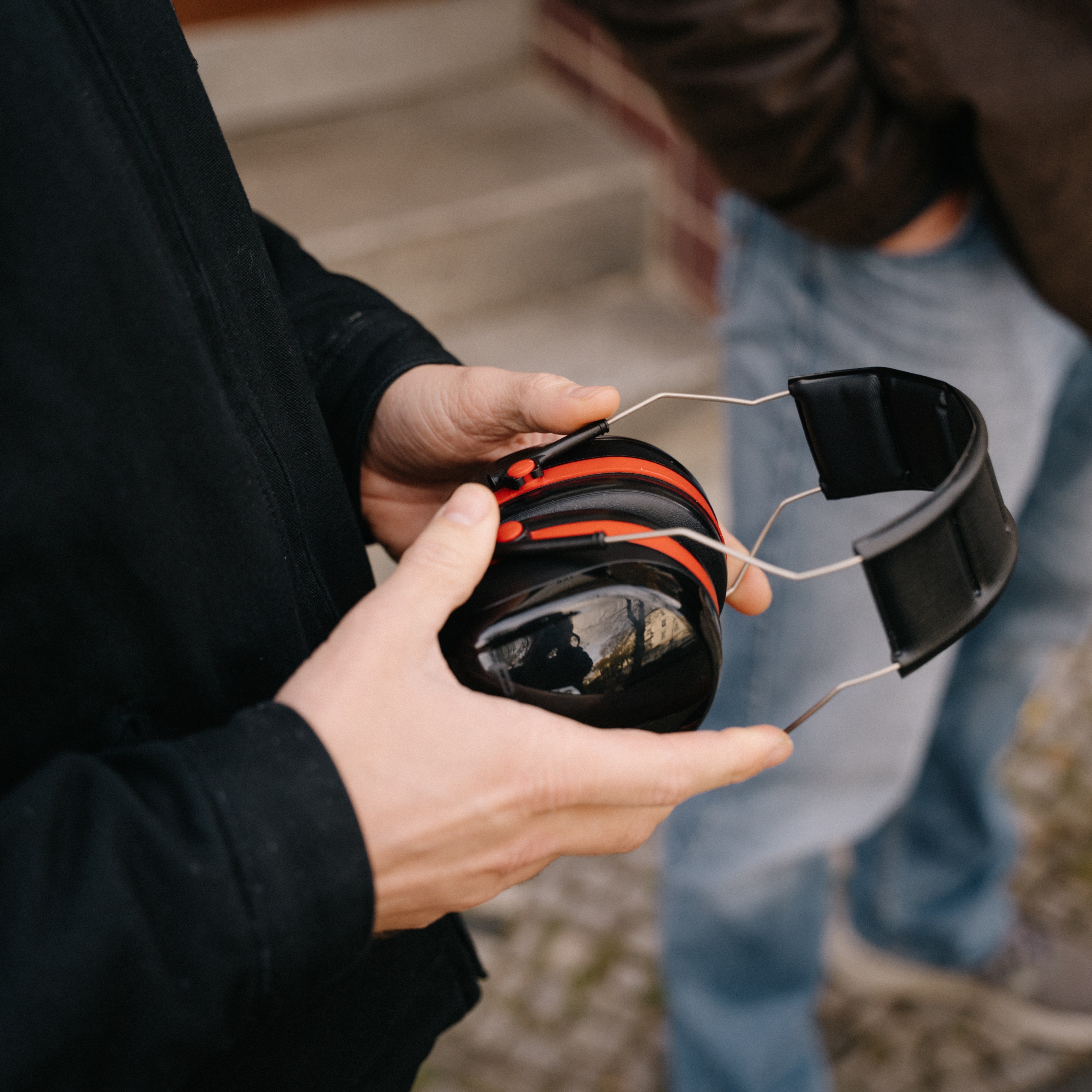 Nahaufnahme der Hände einer Person, die einen schwarz-roten Kapselgehörschutz halten. Im unscharfen Hintergrund steht eine weitere Person in Jeans und brauner Jacke.