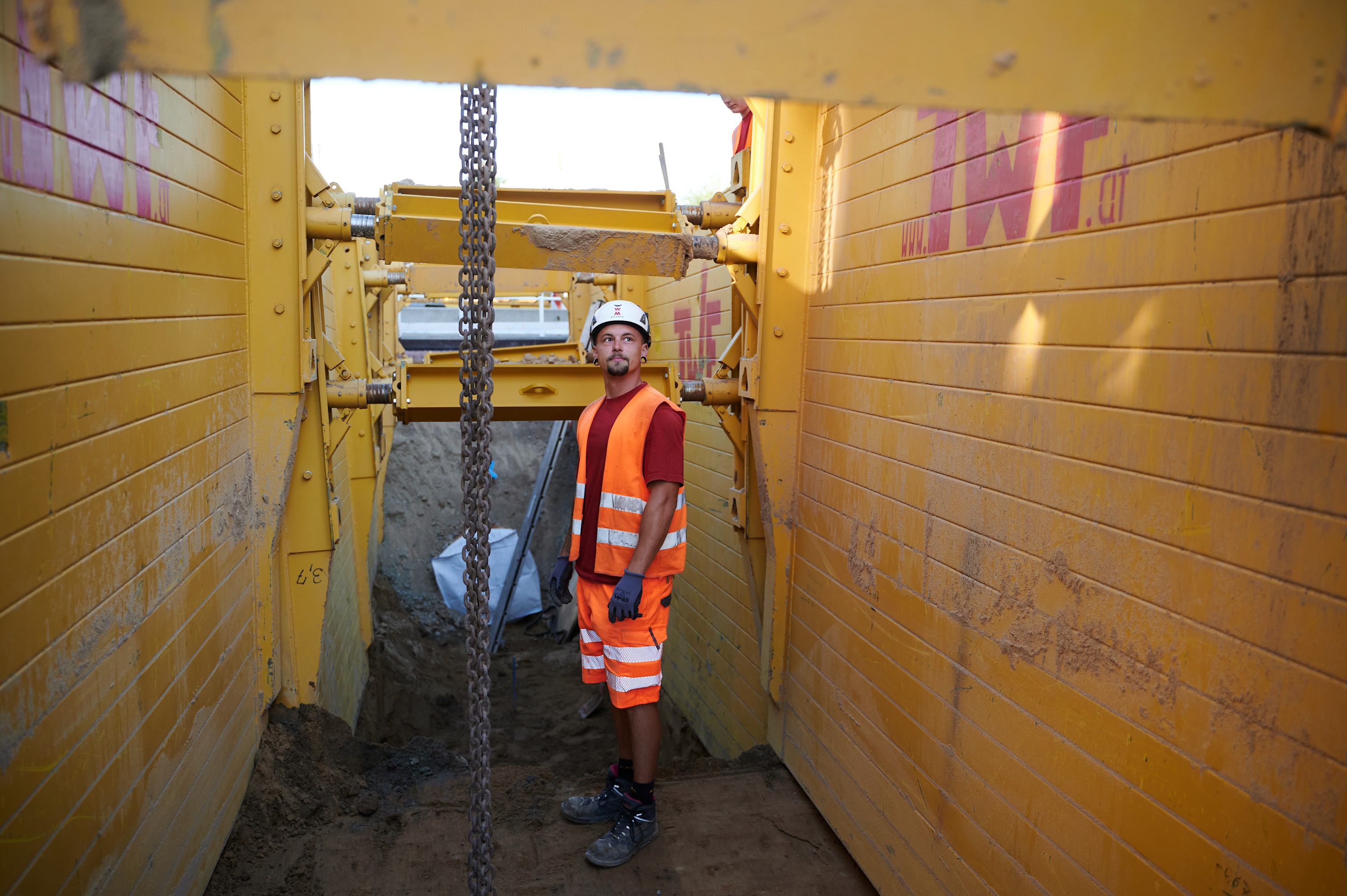 Ein Bauarbeiter in orangefarbener Warnkleidung und Helm steht in einem tiefen Schacht, der mit gelben Stahlplatten gesichert ist. Eine Kette hängt von oben herab.