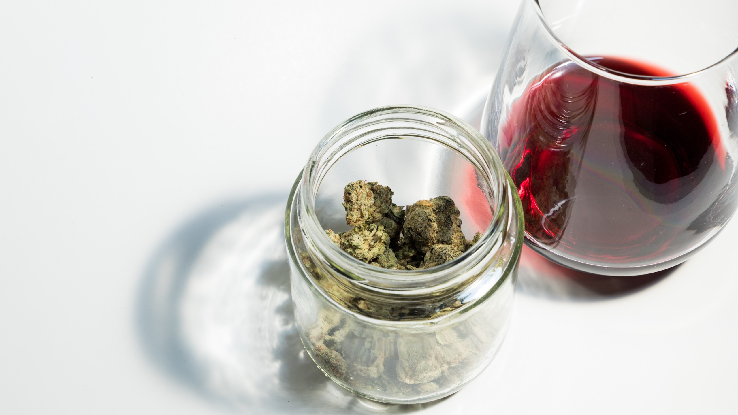 Das Bild zeigt ein kleines, durchsichtiges Glasgefäß, das mit getrockneten Cannabisblüten gefüllt ist. Rechts daneben steht ein Glas, das zur Hälfte mit Rotwein gefüllt ist. Beide Objekte befinden sich auf einer weißen Oberfläche. 