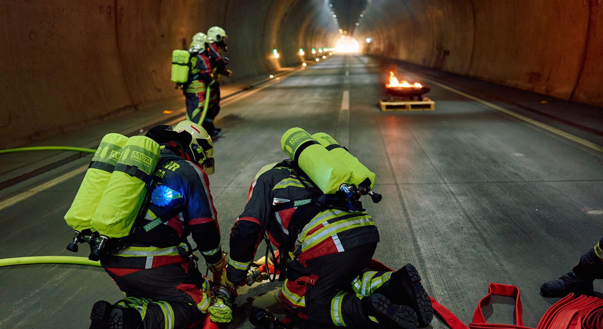 Im Tunnel machen sich Feuerwehrleute bereit: Während im Hintergrund ein Brand lodert, verbinden sie Schläuche für den Löscheinsatz. Ein wichtiger Moment der Sicherheitsübung.