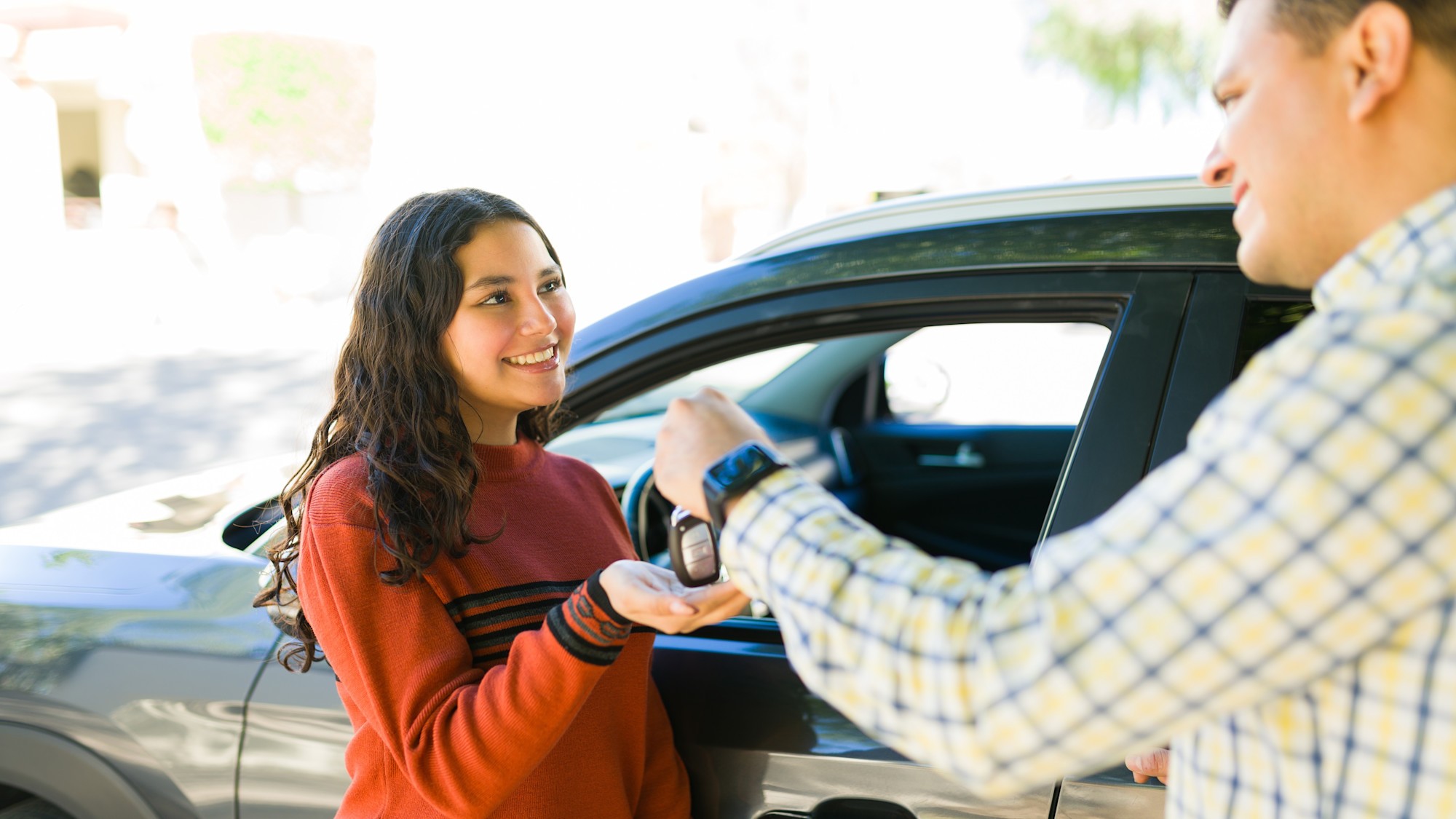 Seitenansicht einer glücklich lächelnden jungen Frau, die von einem Mann einen Autoschlüssel erhält. Sie trägt einen roten Pullover und steht neben einem grauen Auto.