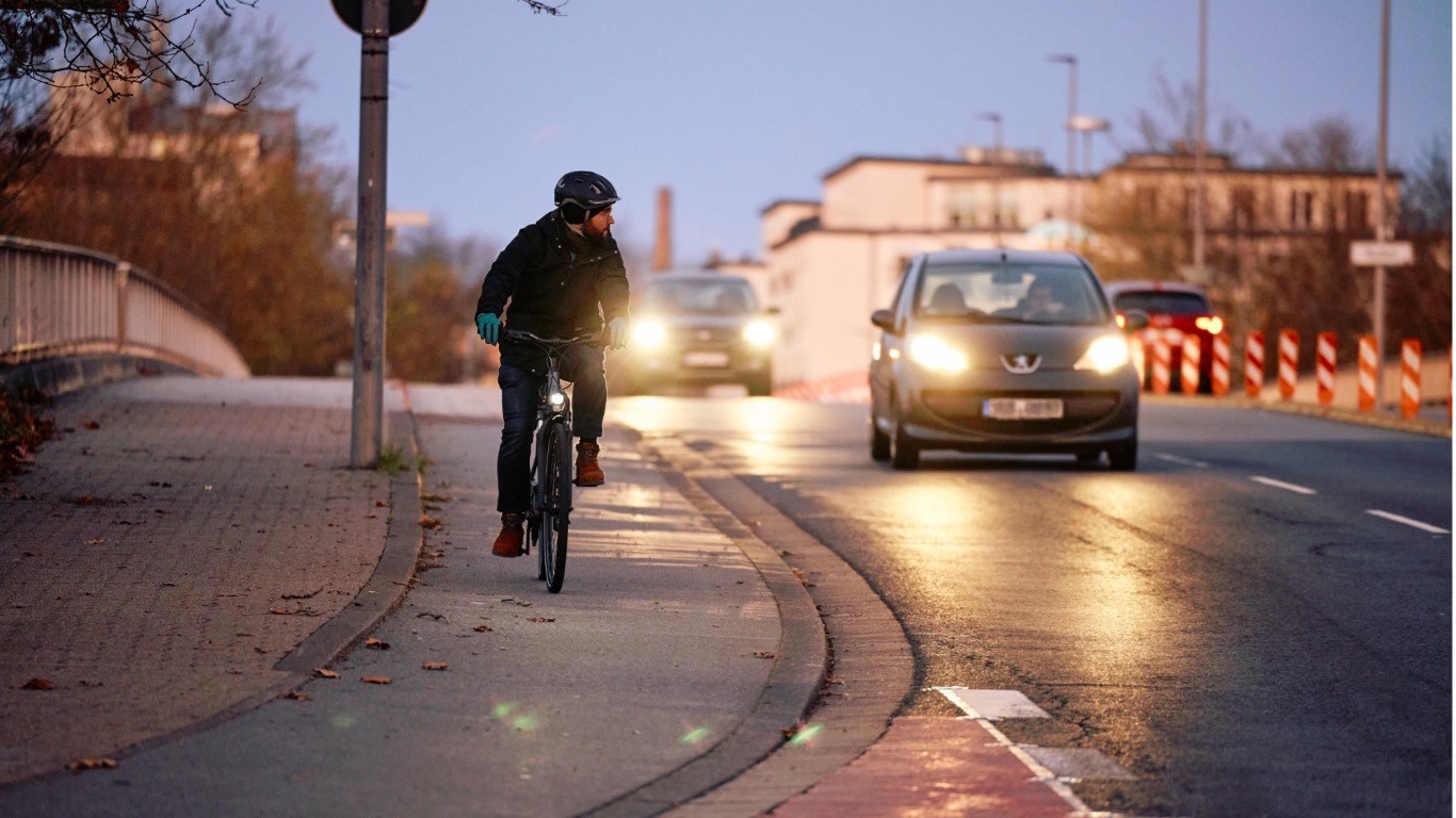 Straße befahren mit Radfahrer und Auto 16:9 Ein Fahrradfahrer fährt bei Dämmerung auf einem Fahrradweg und macht einen Schulterblick. Von hinten nähern sich zwei Autos.