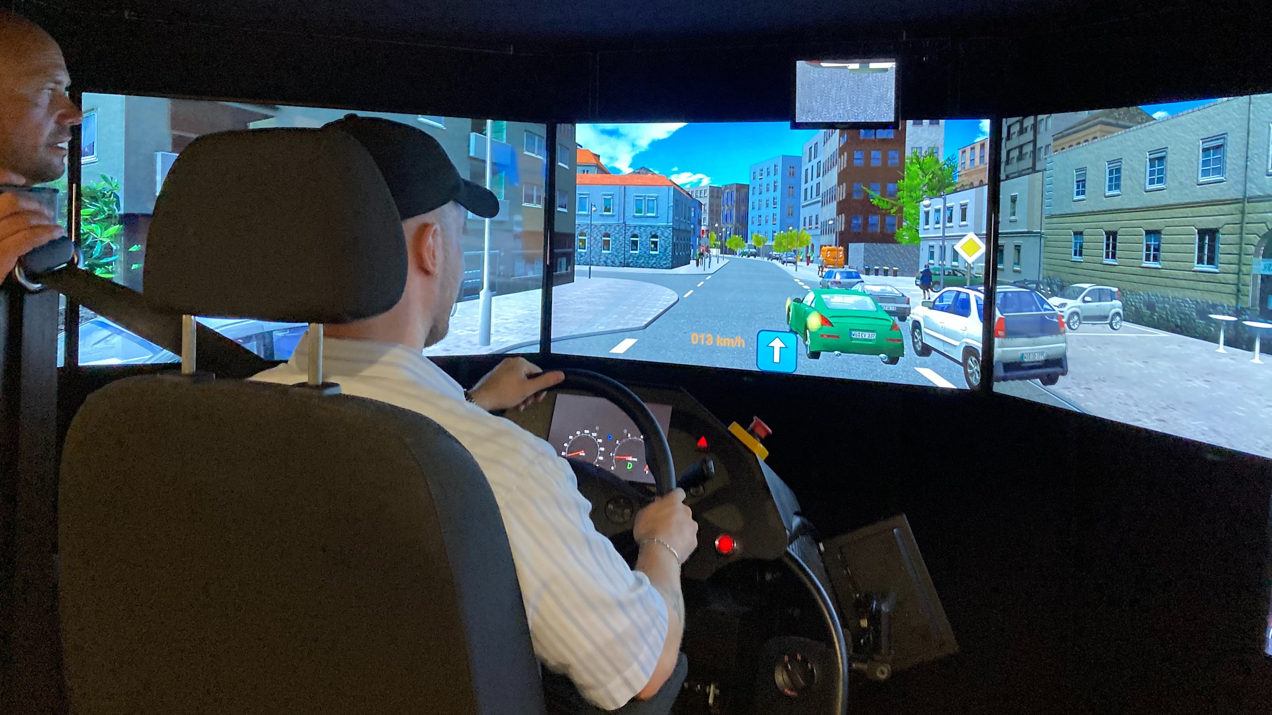 16:9 Einsatzkräfte Verkehrssimulator Das Bild zeigt eine Person in einem Fahrsimulator. Auf mehreren Bildschirmen ist eine virtuelle Stadtstraße zu sehen. Eine weitere Person beobachtet das Geschehen von der Seite.