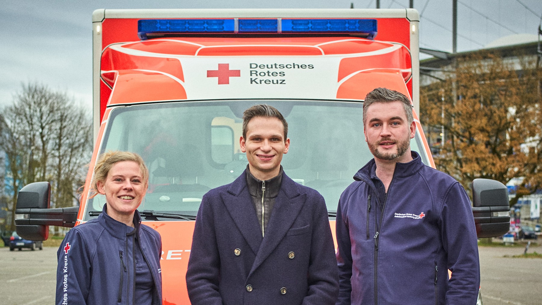 Fabian Köster Polizei Rettungsdienst 16:9 Drei Menschen stehen vor einem parkenden Rettungswagen. In der Mitte steht Fabian Köster.