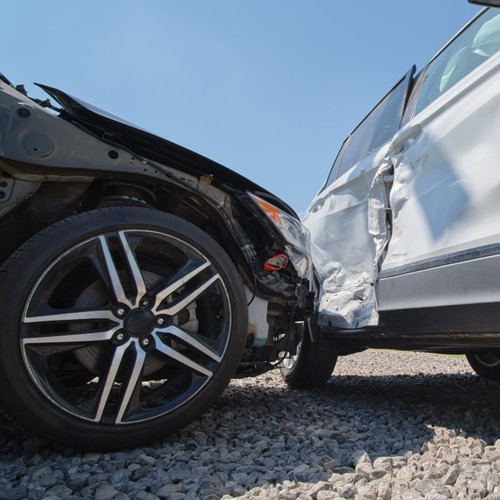 Zu sehen ist eine Nahaufnahme von zwei kollidierten Autos nach einem Unfall: Die Front eines dunklen Autos ist in die Seite eines weißen Autos gefahren. Nach der schweren Kollision sind bei beiden Fahrzeugen erhebliche Blechschäden zu erkennen. 